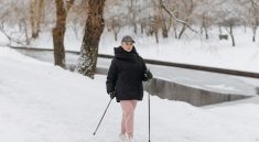 Caucasian elderly woman hiking in winter with trekking poles by a snowy riverbank.