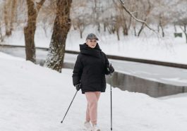 Caucasian elderly woman hiking in winter with trekking poles by a snowy riverbank.